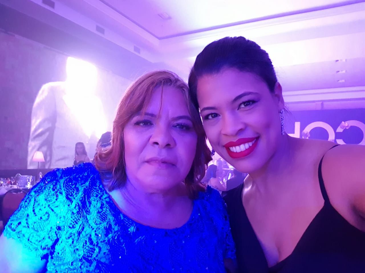 Na foto estão duas mulheres pardas. Da esquerda para a direita está uma mulher de vestido azul e cabelo vermelho, esta é a mãe de Viviane. Ao seu lado está a enfermeira Viviane Dias sorrindo e nos lábios tem um batom vermelho